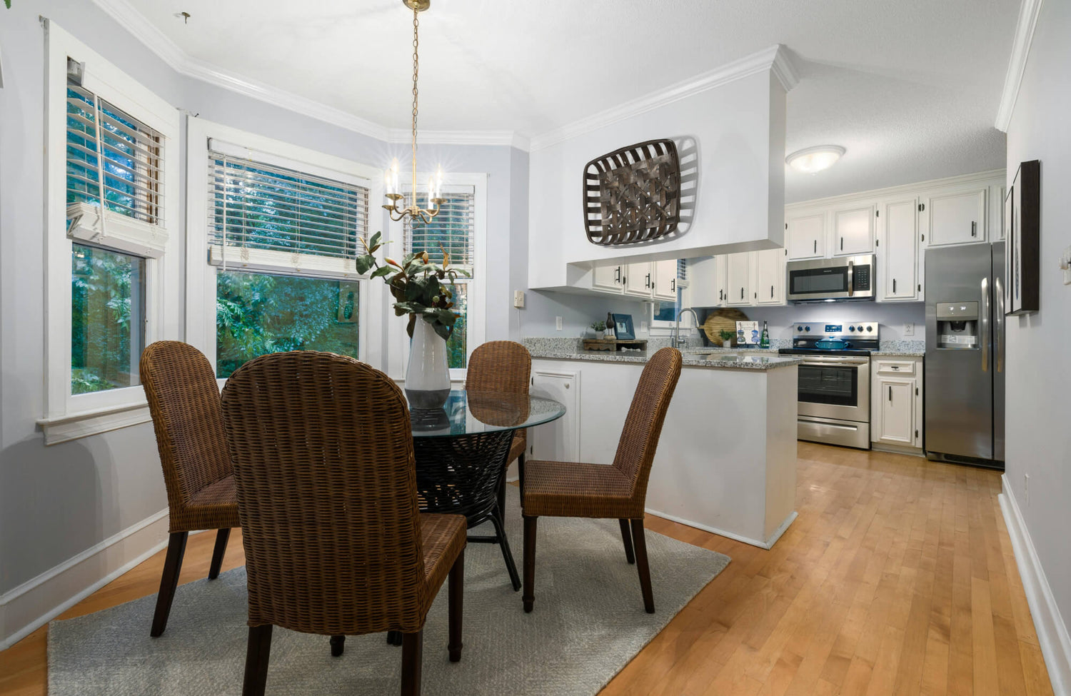Bright dining nook with round glass table, rattan woven chairs, brass chandelier, and adjacent white kitchen with stainless appliances.