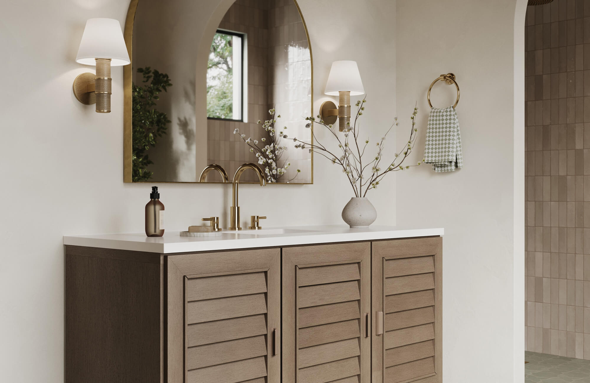 Elegant bathroom vanity with brass fixtures, arched mirror, and wall sconces featuring white shades, styled with branches in a vase.