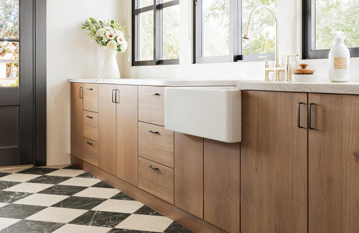 Modern kitchen with wooden cabinets, white farmhouse sink, brass fixtures, and classic black-and-beige checkered marble look flooring.