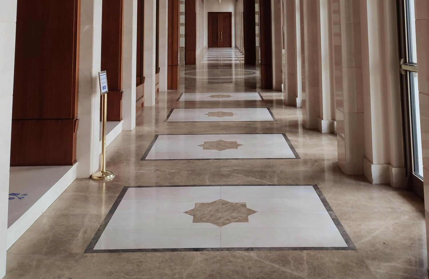 Elegant corridor featuring polished marble flooring with intricate star-shaped inlays, flanked by grand wooden panels and tall columns.