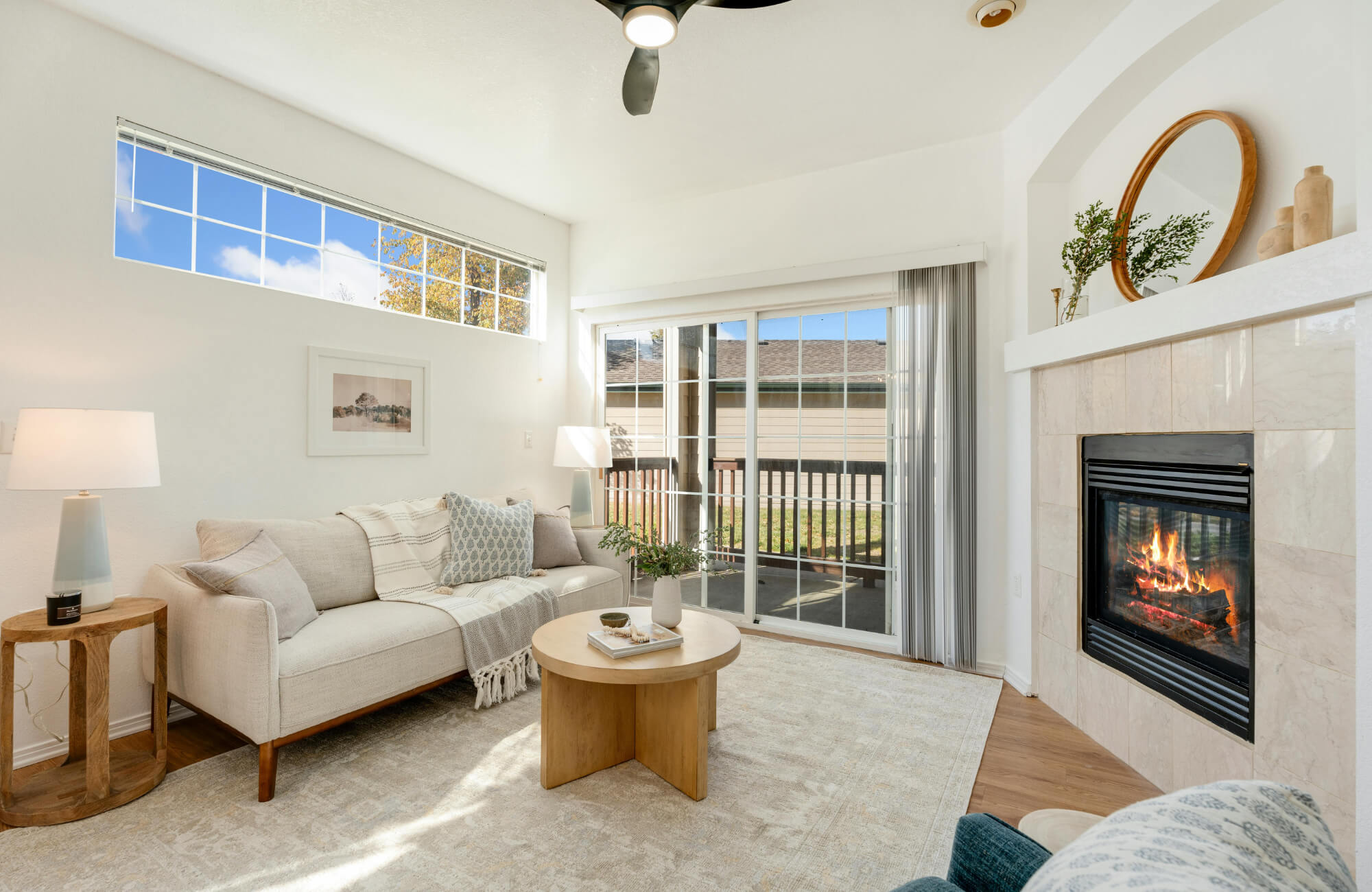 Neutral-toned living room with a light area rug, beige sofa, round wood coffee table, fireplace, and large windows with natural light.