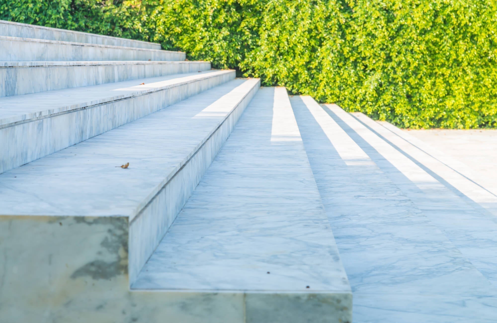 Outdoor marble steps with soft veining, bathed in sunlight and bordered by lush green hedges.
