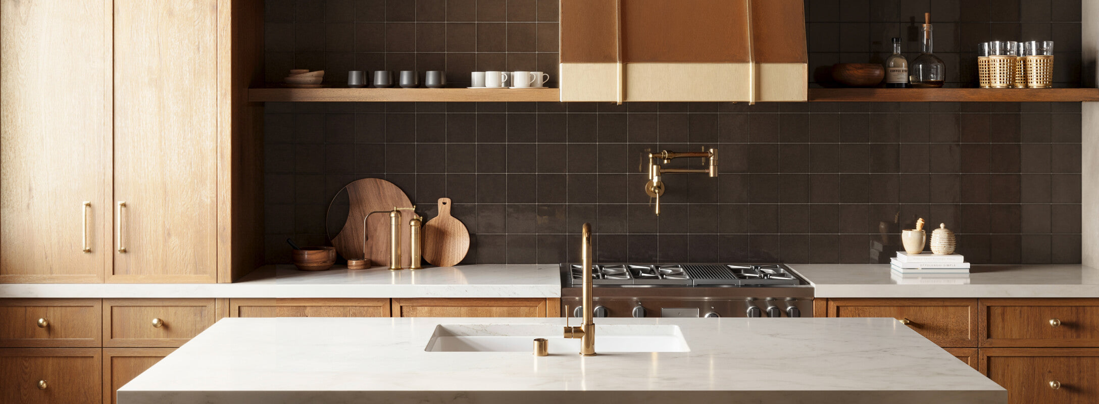 Warm and inviting kitchen with rich brown square backsplash tiles, natural wood cabinetry, and brass fixtures, highlighted by a marble island countertop