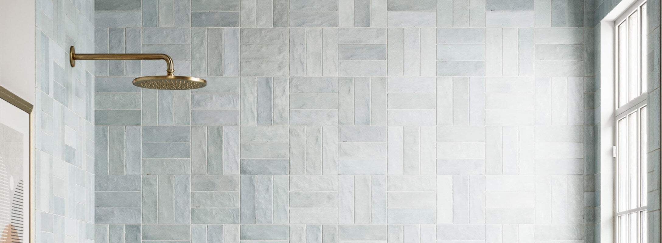 Spacious shower with light blue patterned tiles and a brass showerhead, bathed in natural light from a large window