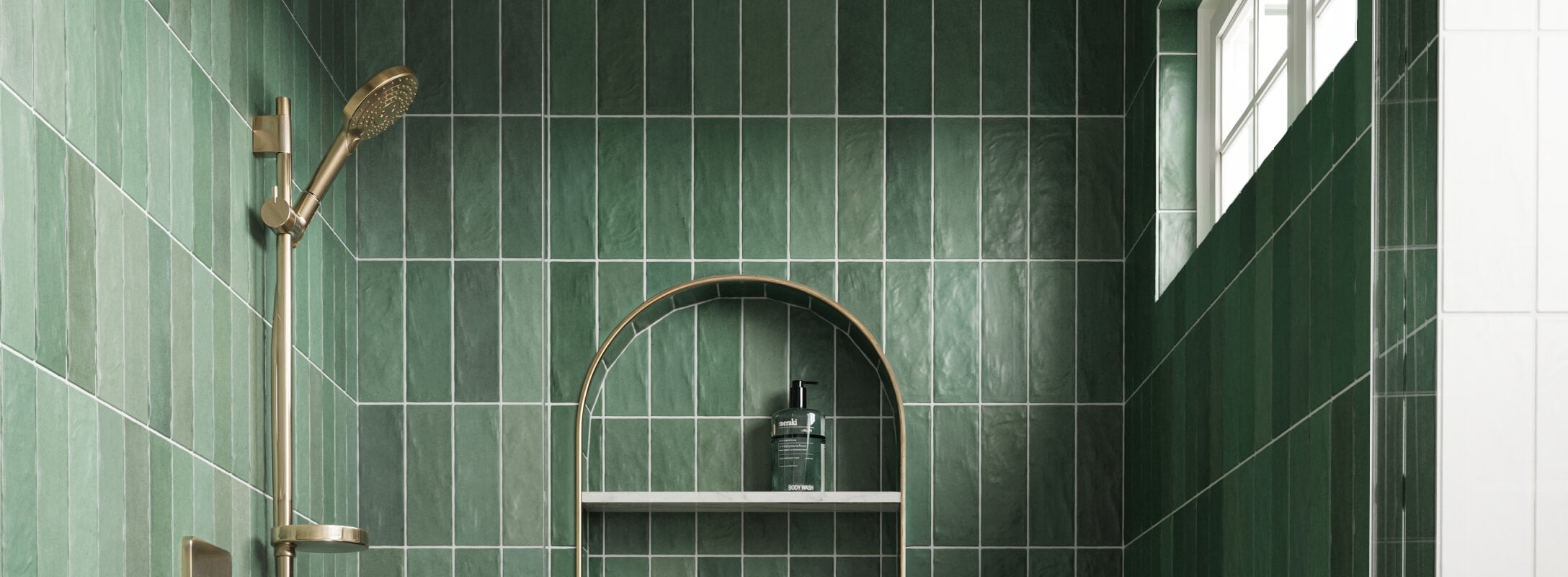 Elegant shower area with vertical emerald green handcrafted look tiles, a brass showerhead, and an arched niche holding body wash, illuminated by natural light from a window