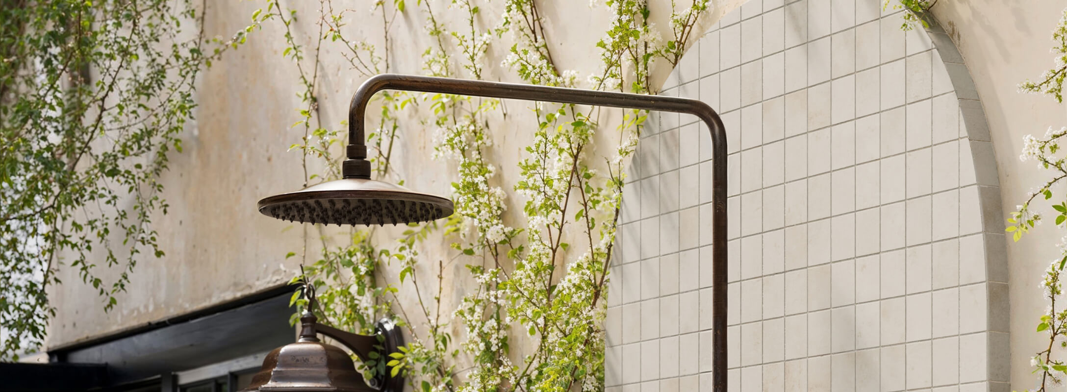 Outdoor shower with taupe mosaic tiles on the wall, rustic brass showerhead, and lush greenery, creating a natural and serene setting