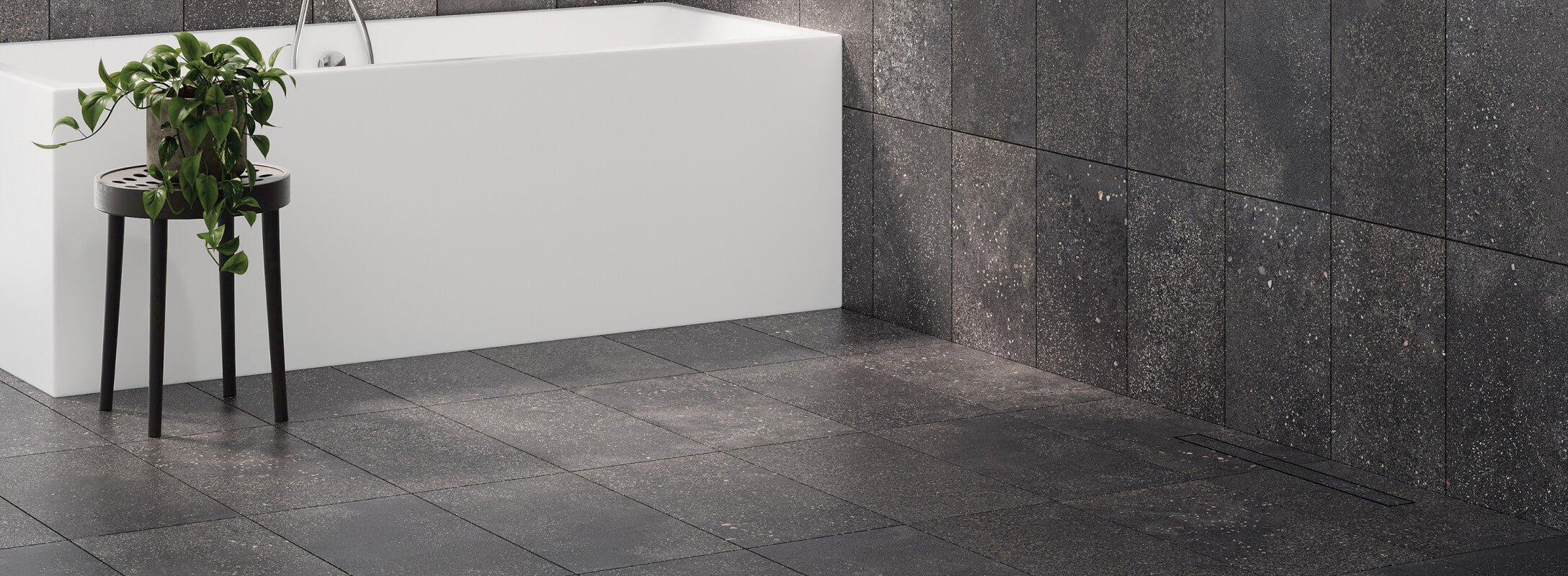 Dark grey terrazzo look floor tiles complement a modern white bathtub, with a lush plant adding contrast