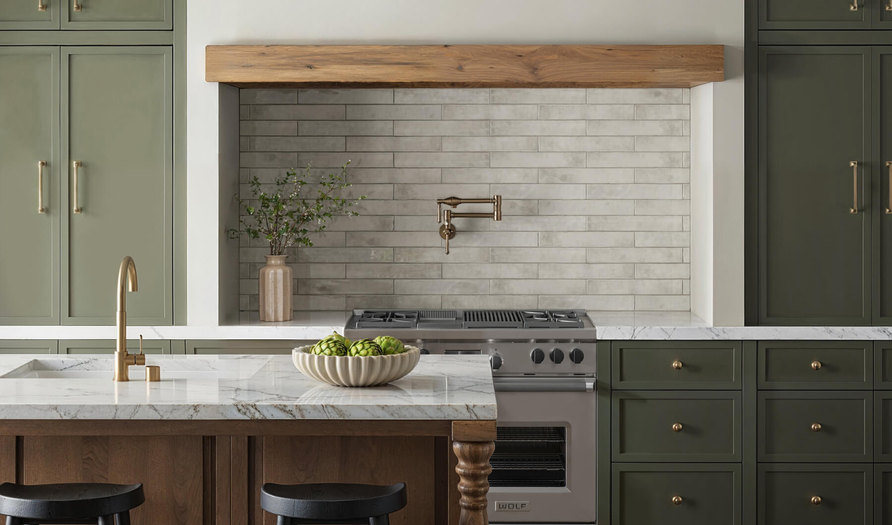Elegant kitchen with light grey subway tiles, olive green cabinetry, and brass fixtures, complemented by a white marble countertop and a wooden hood