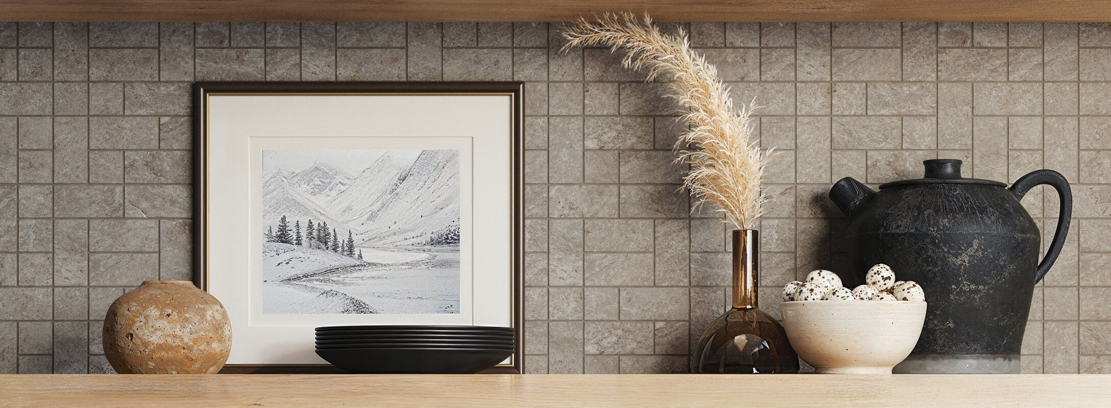 Taupe mosaic tiles create a rustic backsplash, complemented by earthy decor including pottery, a framed landscape, and dried pampas grass