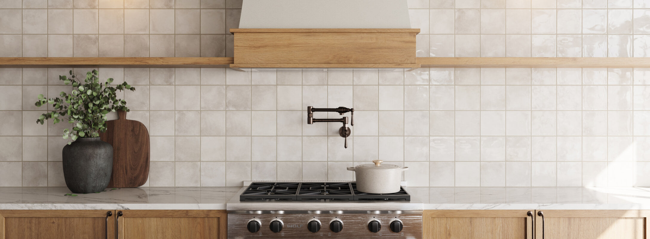 Modern kitchen with beige decorative tiles, light wood cabinetry, and a central range hood. The countertop features a marble surface with a dark vase and greenery for added decor