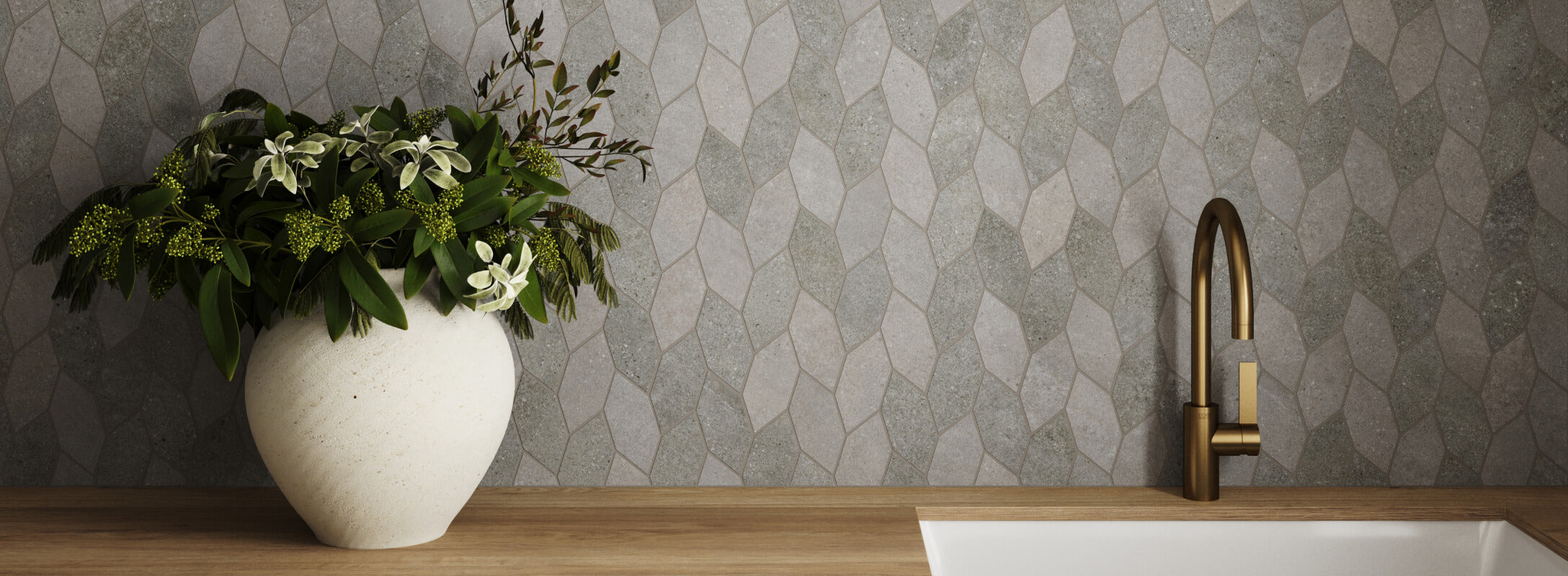 Geometric grey wall tiles create a textured backdrop for a minimalist sink, brass faucet, and a large vase filled with lush greenery