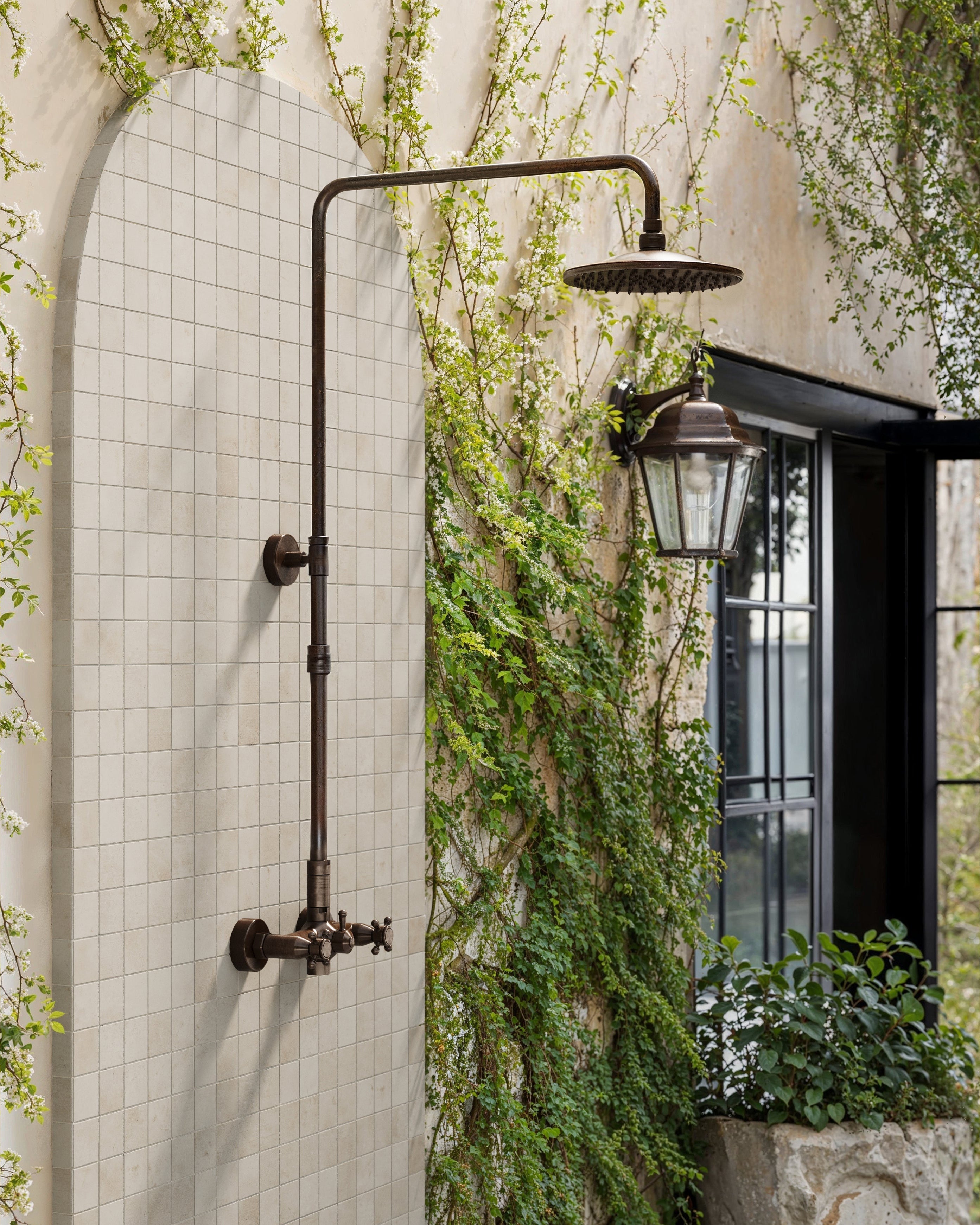Rustic outdoor shower with matte 2x2 taupe mosaic tiles, framed by lush green vines and a vintage lantern