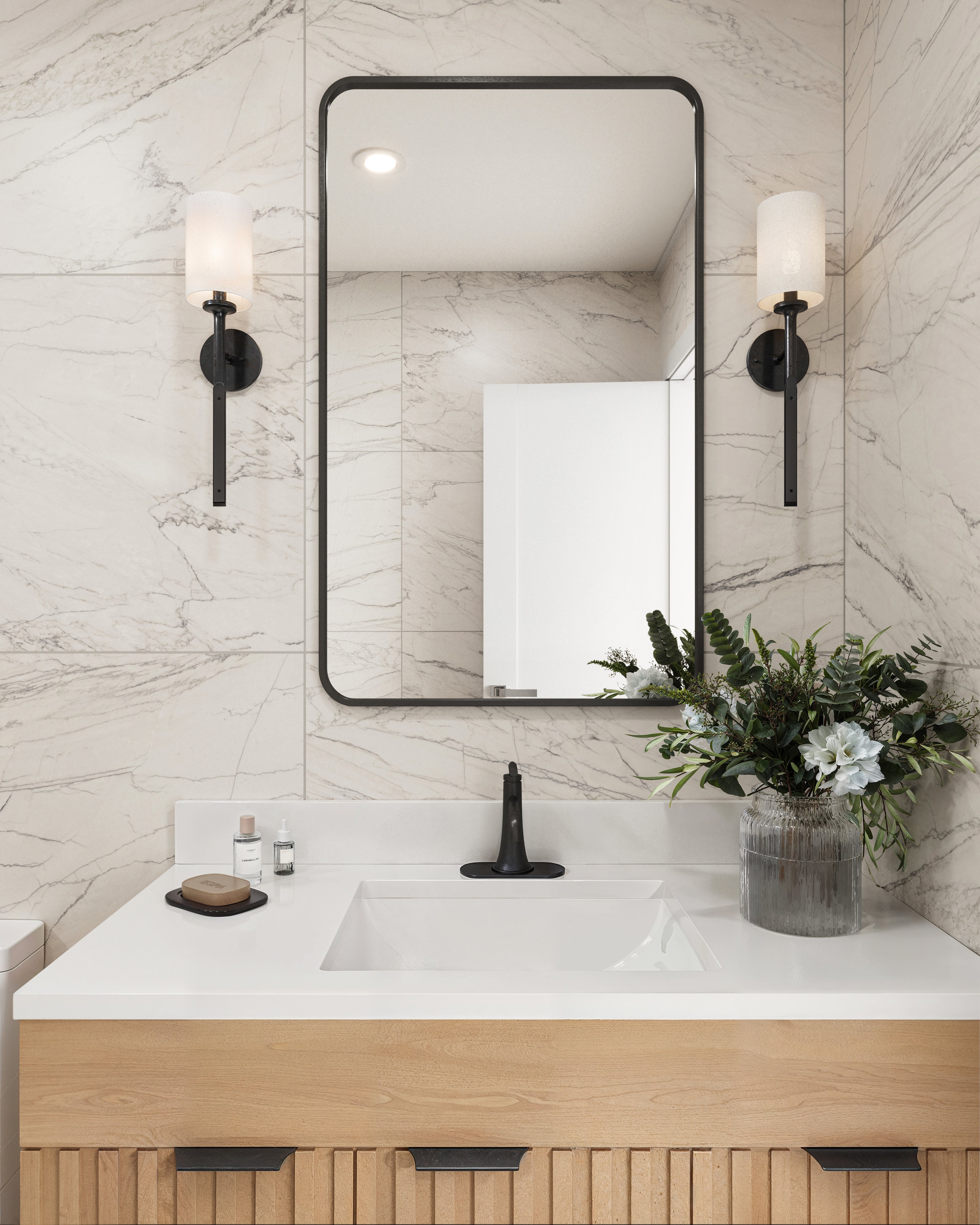 A modern vanity area featuring marble look walls, a light wood fluted cabinet, black fixtures, and sleek sconces, accented with fresh greenery for a polished aesthetic.