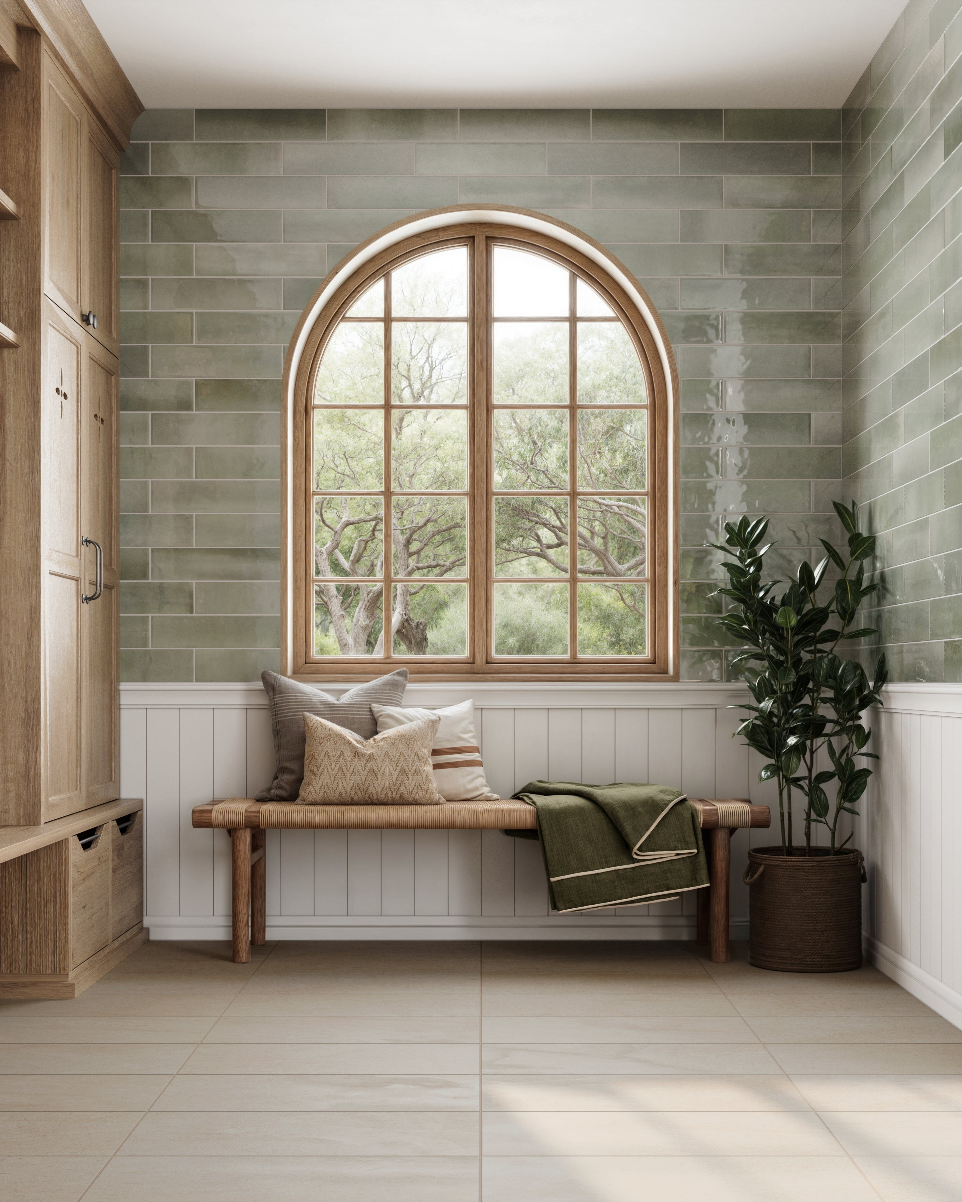 A cozy mudroom featuring glossy green subway tiles, a natural wood bench with soft pillows, and an arched window overlooking lush greenery.