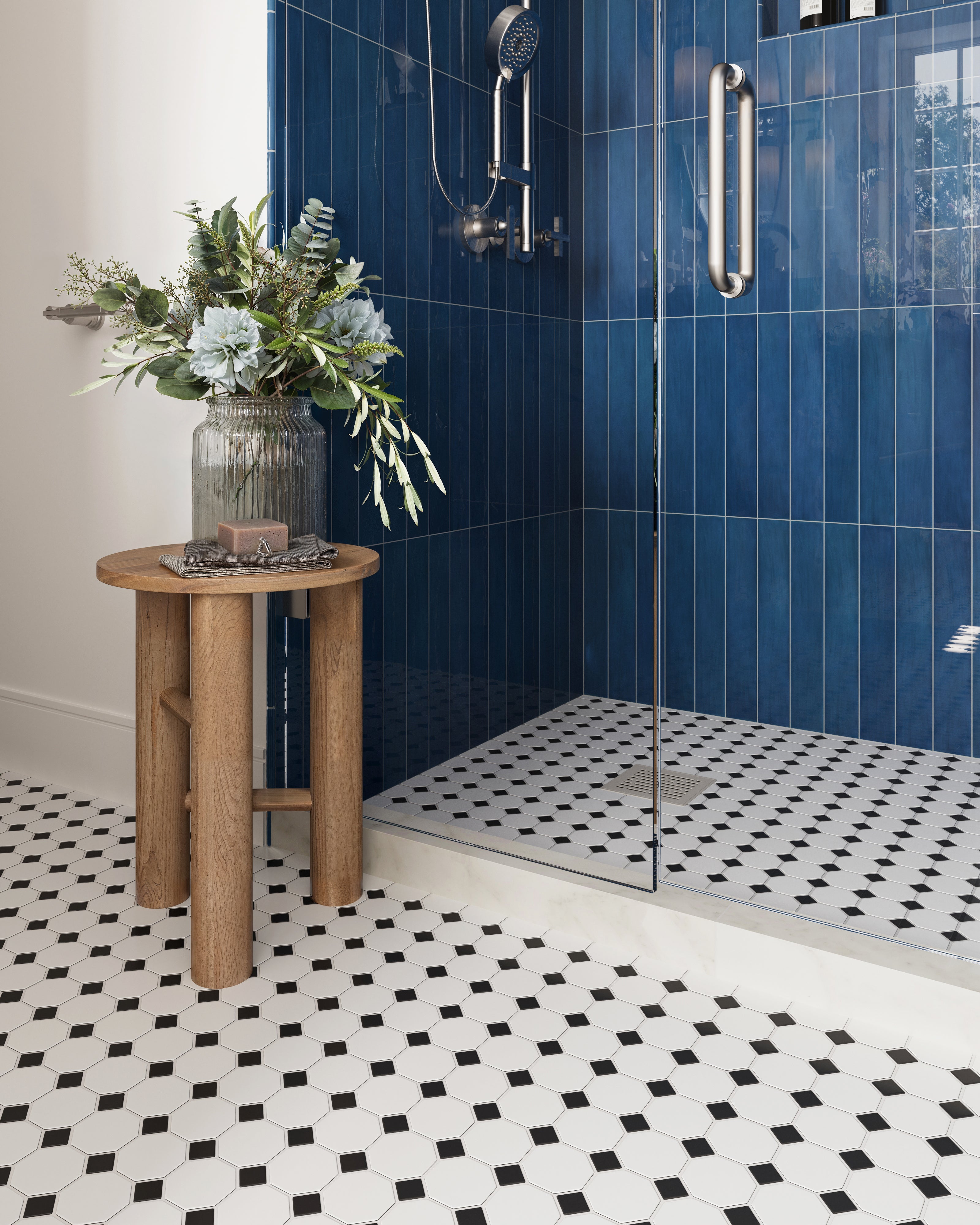 Chic bathroom with glossy blue shower tiles, a black-and-white octagon mosaic floor, and a rustic wood side table holding fresh greenery.