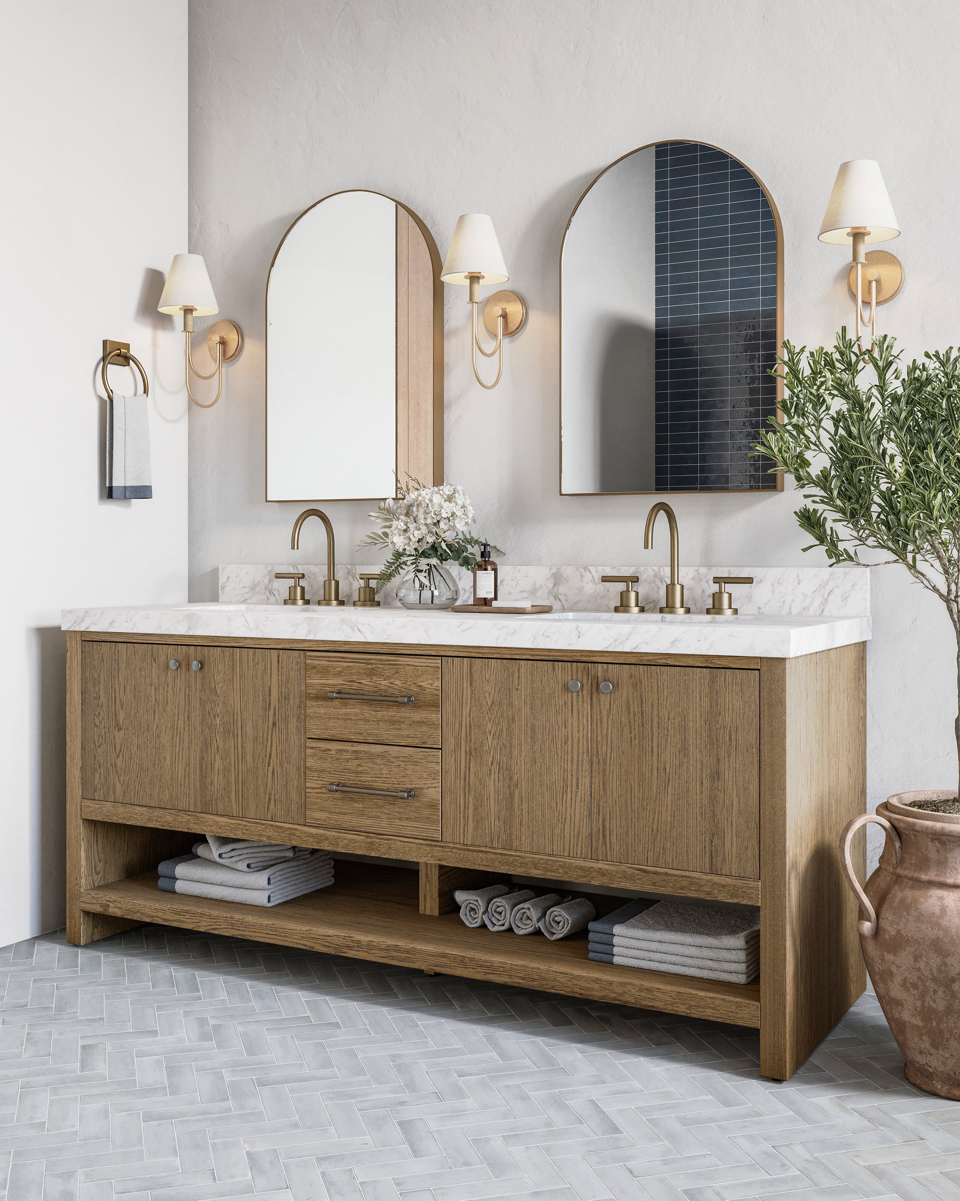 Two arched mirrors with brass frames are mounted above a marble double vanity with wood cabinetry and gold wall sconces in a bright bathroom.