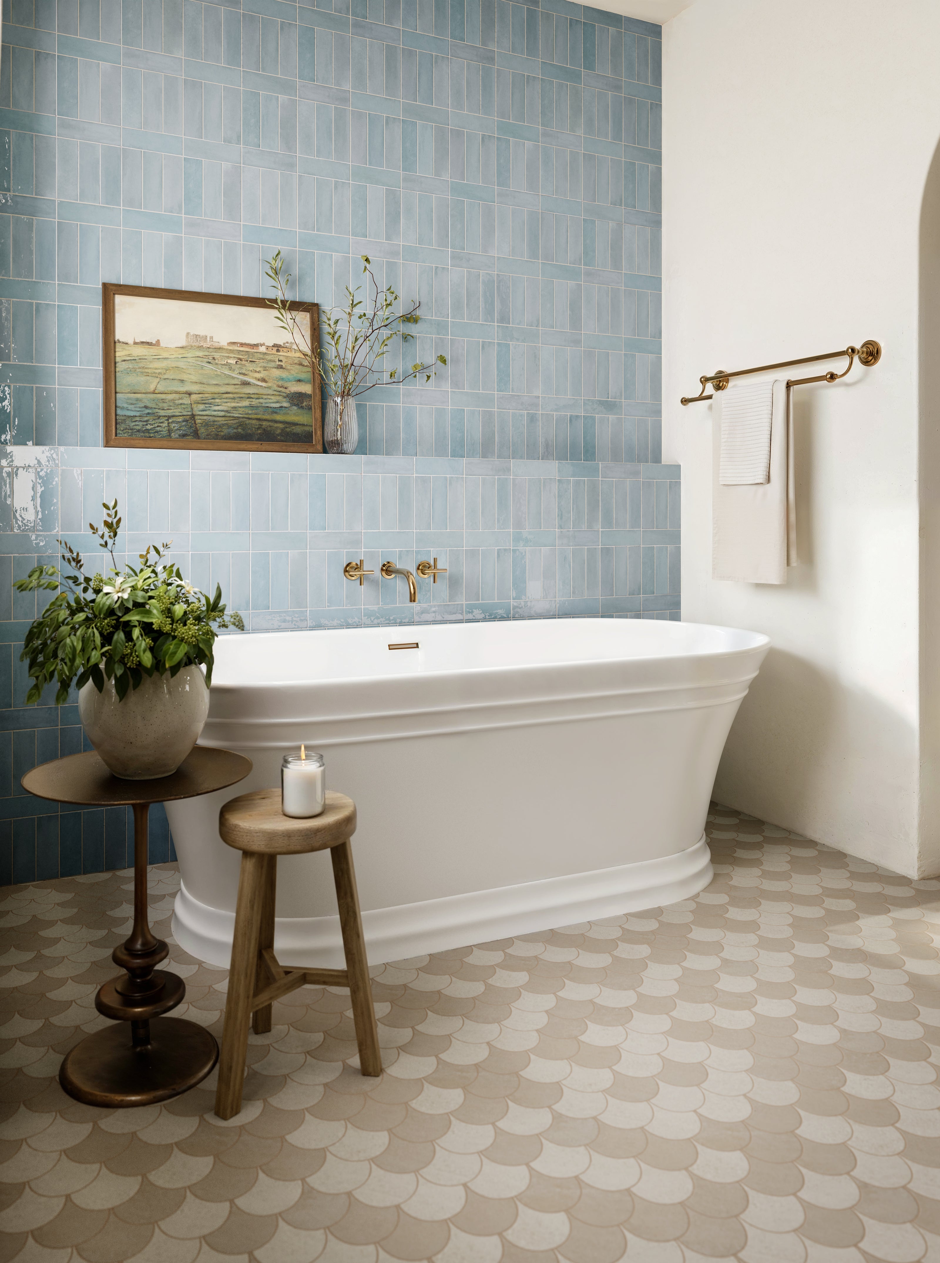 Bathroom with a white freestanding tub against a wall of glossy pastel blue subway tiles with rounded edge trim, gold fixtures, scalloped beige floor tiles, and a wooden side table with a plant and candle