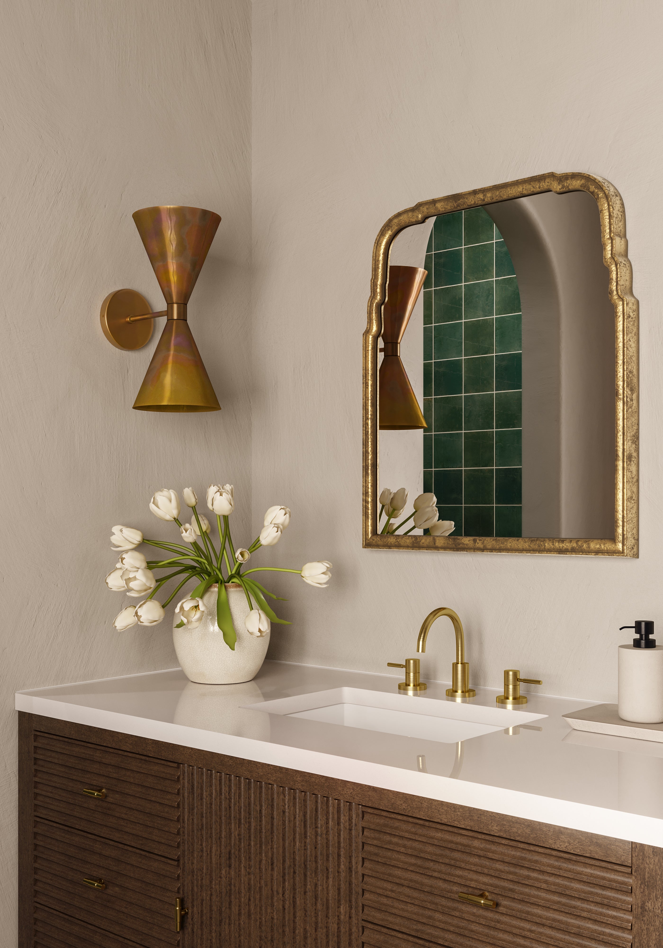 Wood vanity with white countertop, brass faucet, gold framed mirror, sculptural brass sconce, and vase of white tulips against soft walls