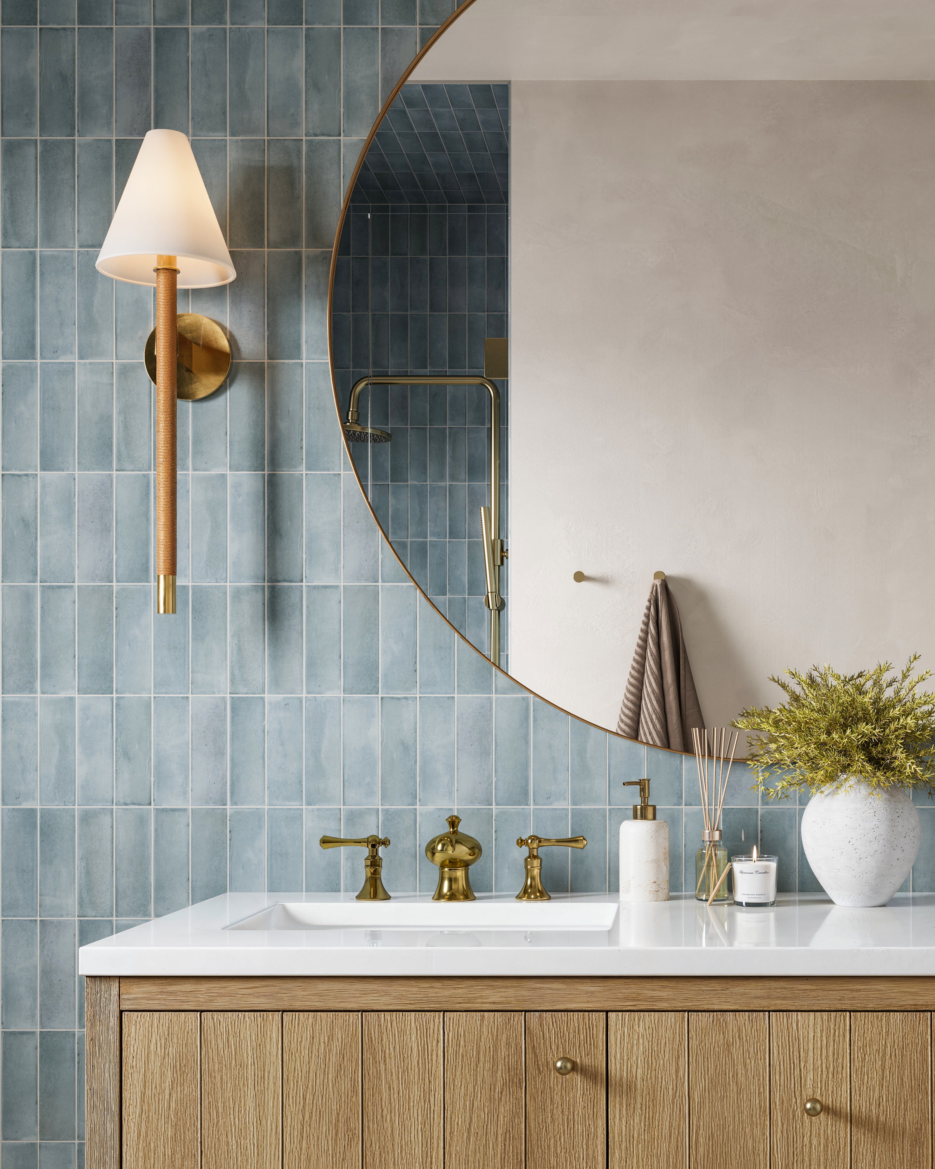 Detail of round polished brass mirror, aged brass faucet, and oak vanity set against vertical blue tile.