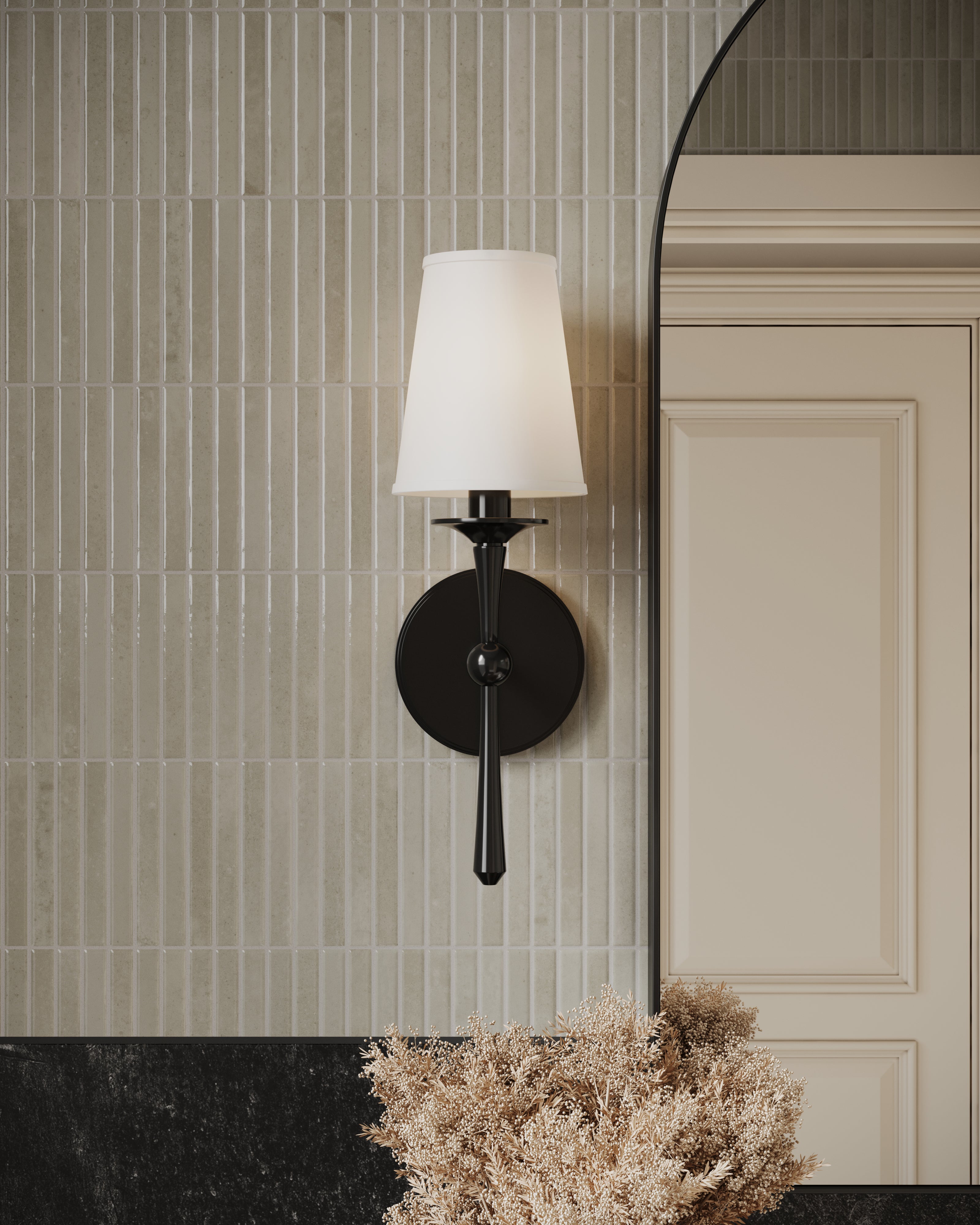 Matte black wall sconce with tapered white shade mounted on vertical ceramic tile, beside an arched mirror and natural florals