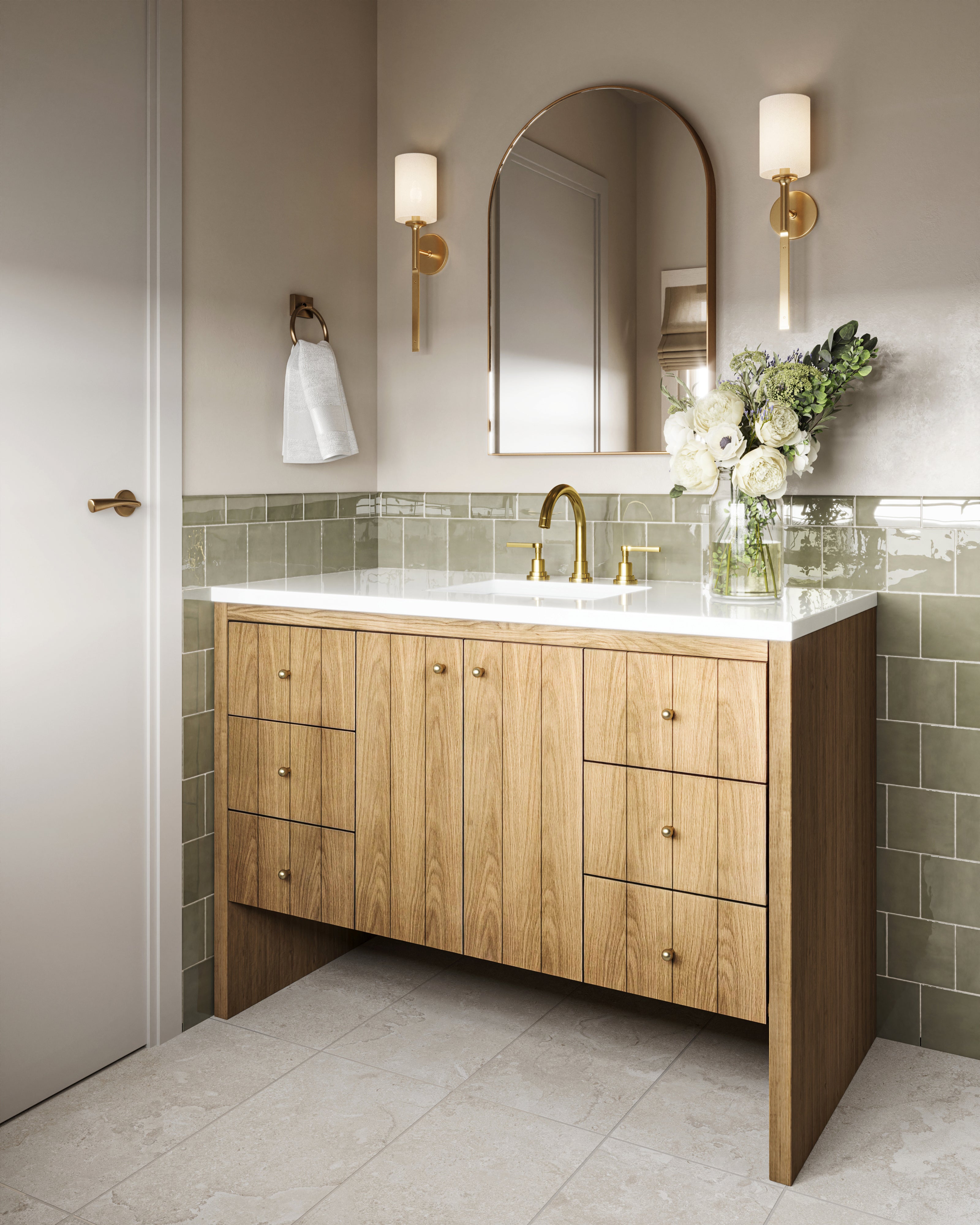Light natural oak bathroom vanity with vertical wood grain doors, side drawers with brass knobs, and a white quartz countertop with integrated sink and brass faucet. Styled with an arched mirror, brass wall sconces, soft green tile backsplash, and a vase of white flowers on the counter.