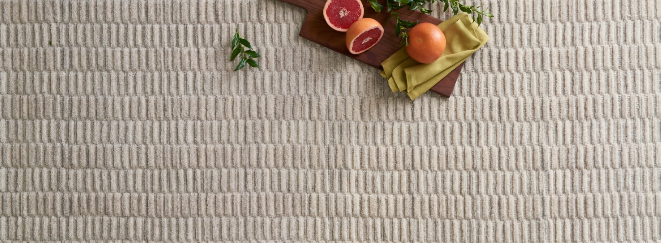 Hand loomed area rug in soft beige tones with a raised linear texture, styled with fresh citrus and greenery for a serene aesthetic.