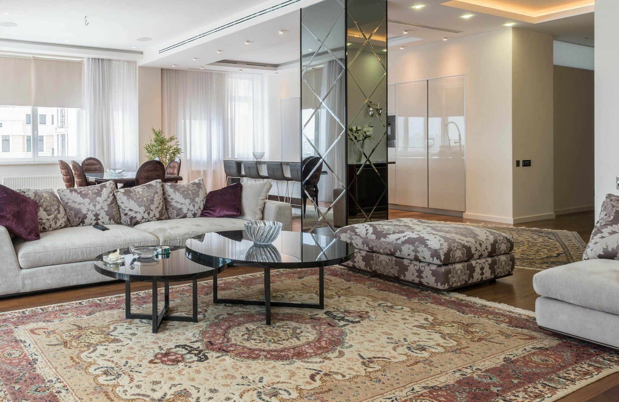 Elegant living room with a large 9x12 rug featuring intricate floral patterns in warm beige and red tones, creating a refined foundation for the modern sectional sofa and glass-top tables.