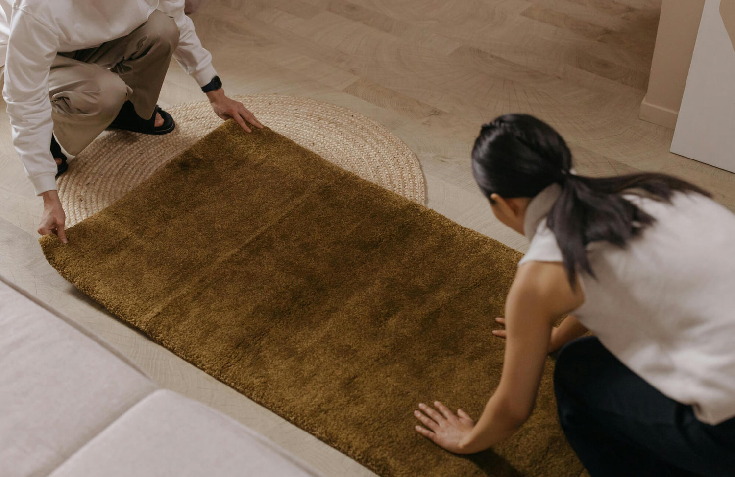 Two people are arranging a brown area rug over a woven mat in a cozy living room with light wood flooring.