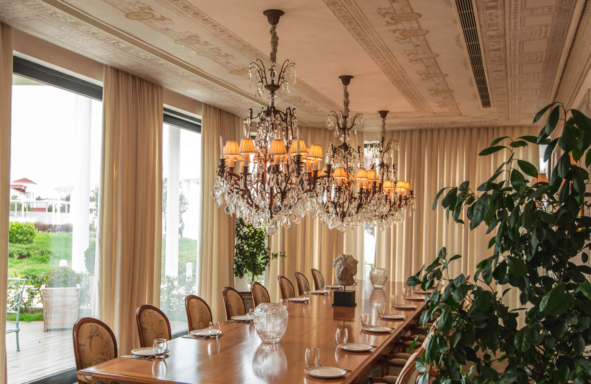 Elegant dining room with a long polished wood table, ornate crystal chandeliers, and tall windows draped in cream curtains.