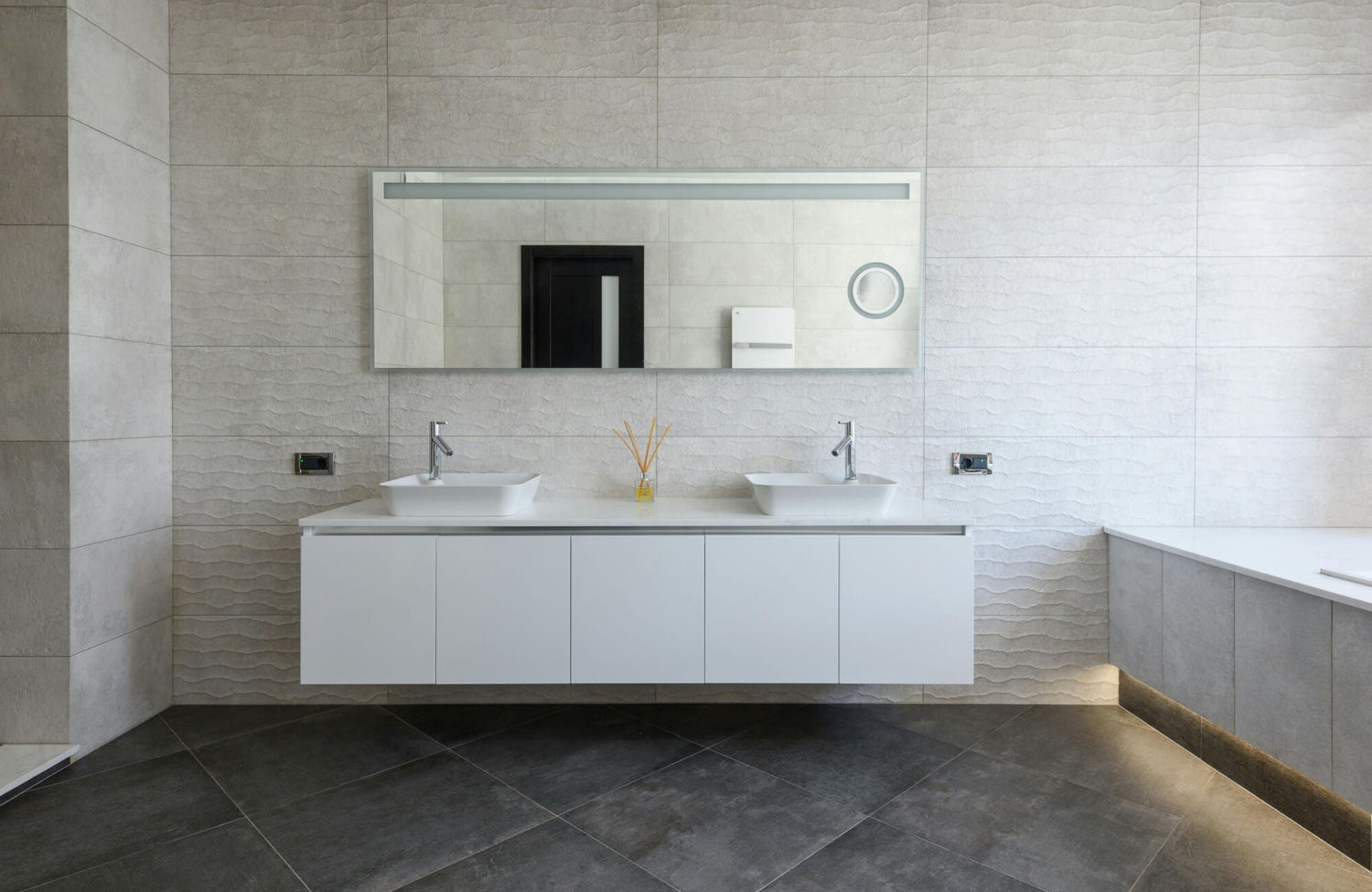 Contemporary bathroom vanity with a floating white cabinet, dual vessel sinks, and sleek chrome faucets set against textured neutral wall tiles, creating a clean, balanced, and modern bathroom aesthetic.