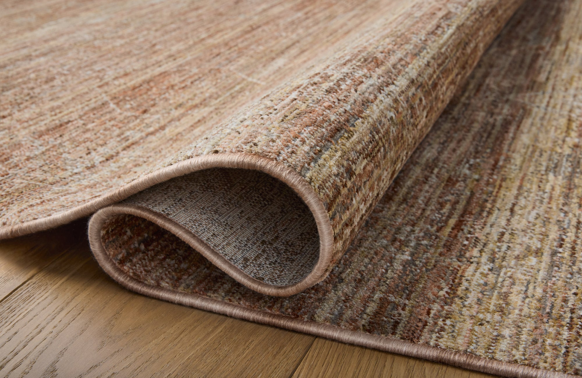 Close-up of a rolled rug showing its dense weave and multi-tonal color pattern on a hardwood floor.