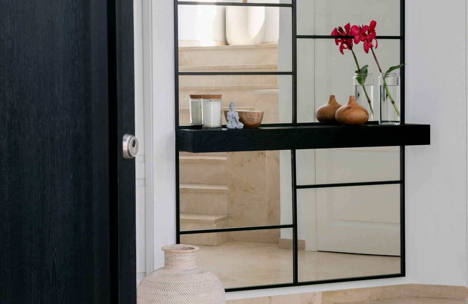 A modern entryway featuring a large floor mirror with black grid-style framing, reflecting the neutral stone staircase and styled with minimalist décor including candles, wooden accents, and fresh florals for a clean, contemporary look.