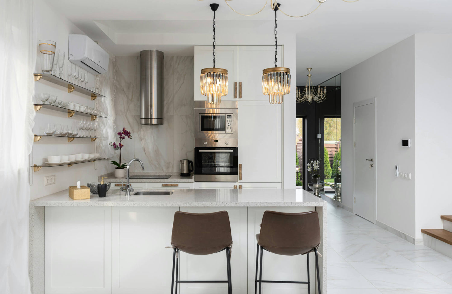 Two modern counter stools with slim black metal legs and curved brown upholstered seats are positioned at a bright white kitchen island, complementing the clean cabinetry, marble-look backsplash, and warm brass lighting.