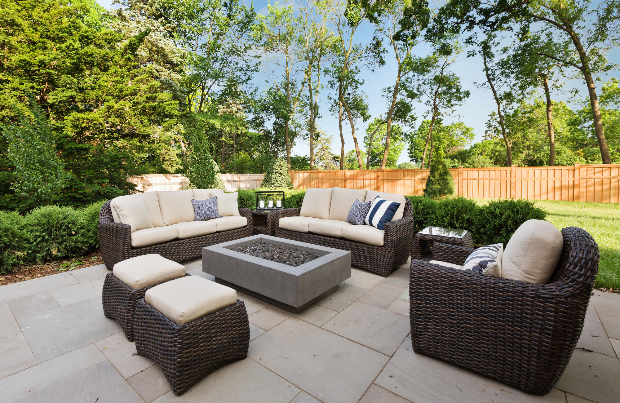 Outdoor patio with wicker furniture, beige cushions, a modern fire pit, and large stone tiles surrounded by lush greenery and a wooden fence.