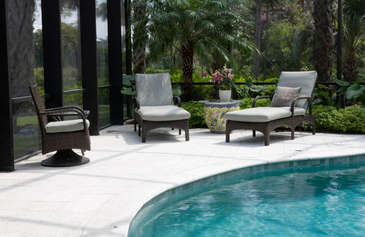 Poolside lounge area with wicker chairs and white non-slip tile flooring, surrounded by lush greenery and enclosed by glass panels.