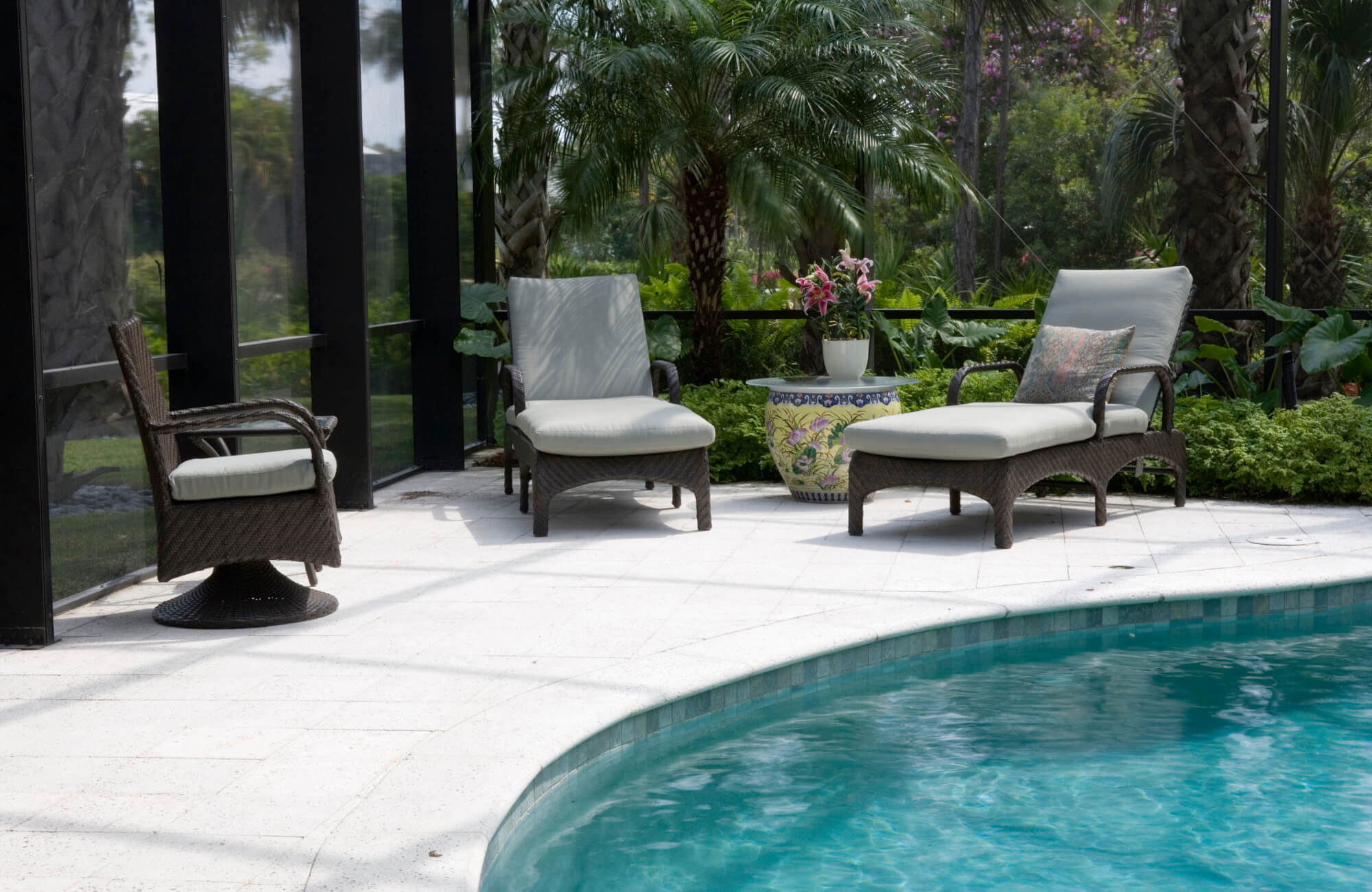 Poolside lounge area with wicker chairs and white non-slip tile flooring, surrounded by lush greenery and enclosed by glass panels.