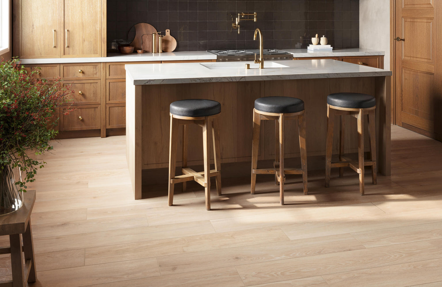 Warm kitchen with brown wood cabinetry, white countertops, a dark brown tile backsplash, and light brown wood look tile flooring beneath a central island with bar stools.
