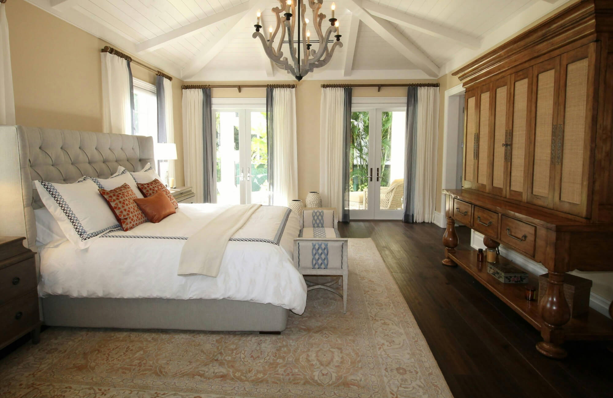 Elegant bedroom with a wool rug, a tufted upholstered bed, warm wood furnishings, and large windows opening to a lush garden view.