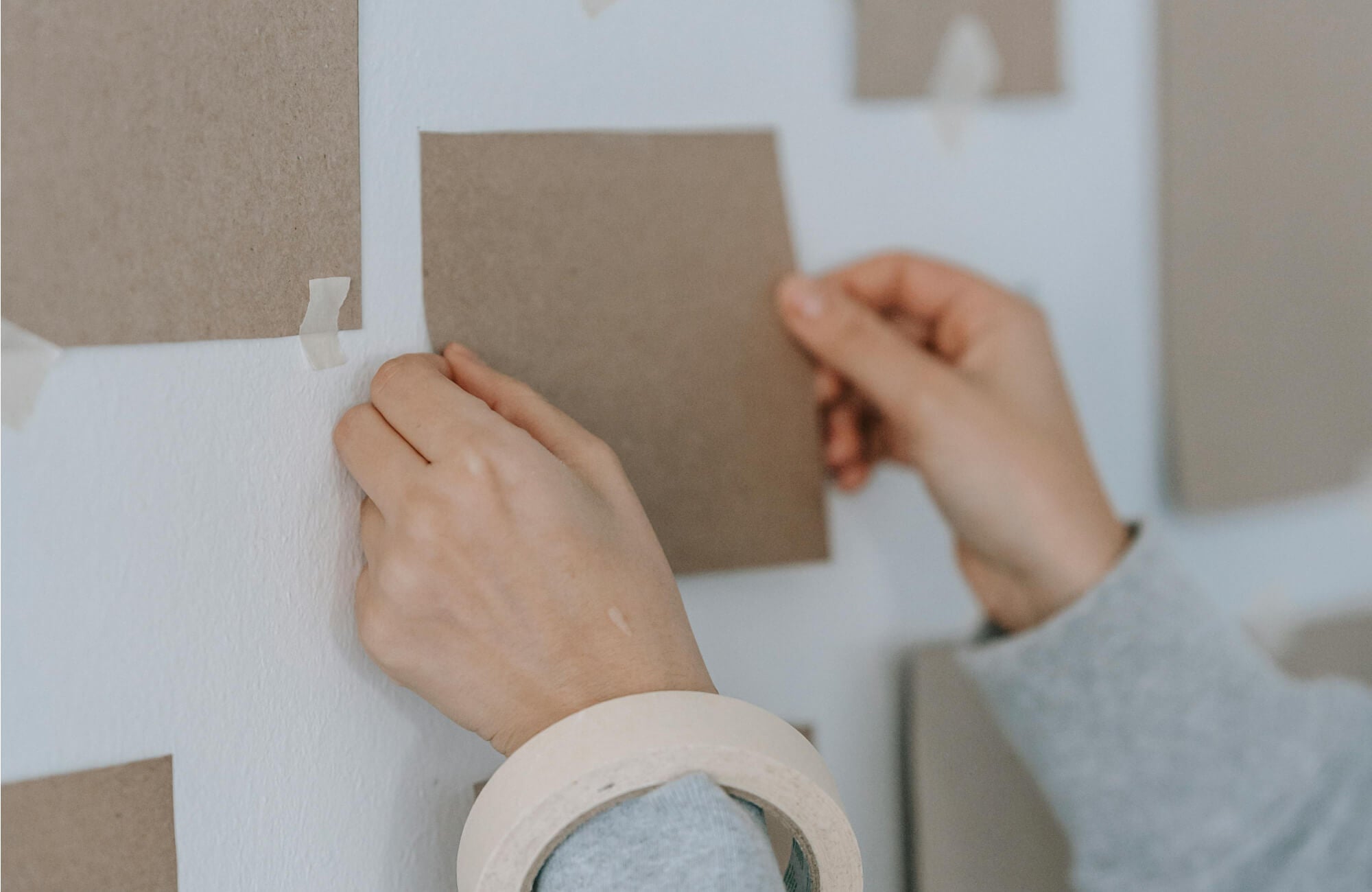 DIY wall art setup with hands taping neutral brown paper pieces onto a white wall for a minimalist gallery arrangement.