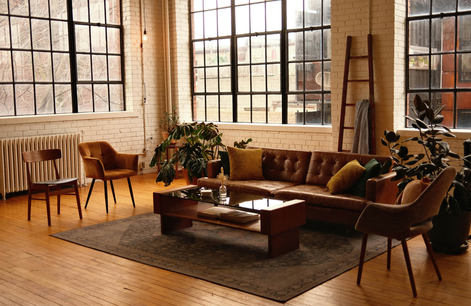 Industrial-style loft living room with large windows, mid-century furniture, and a textured brown rug grounding the cozy seating area.