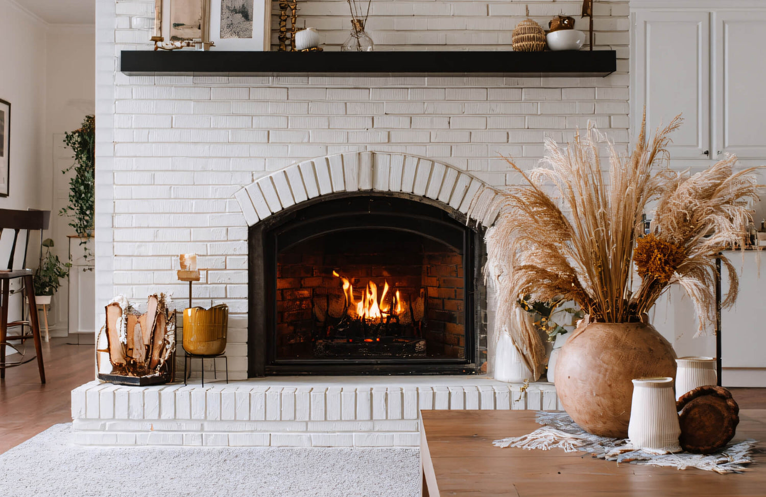 White-painted brick fireplace with a black mantel, warm fire, and neutral modern décor including dried grasses, pottery, and wood accents.