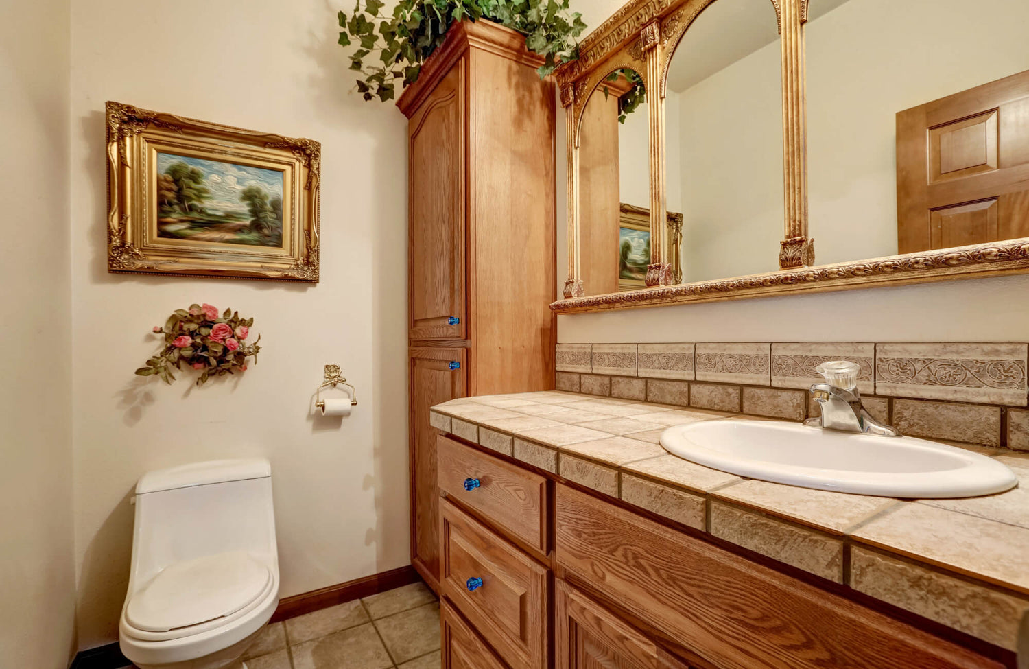 Traditional bathroom with a wood vanity, tiled countertop, ornate framed mirror, wall art, and decorative greenery.
