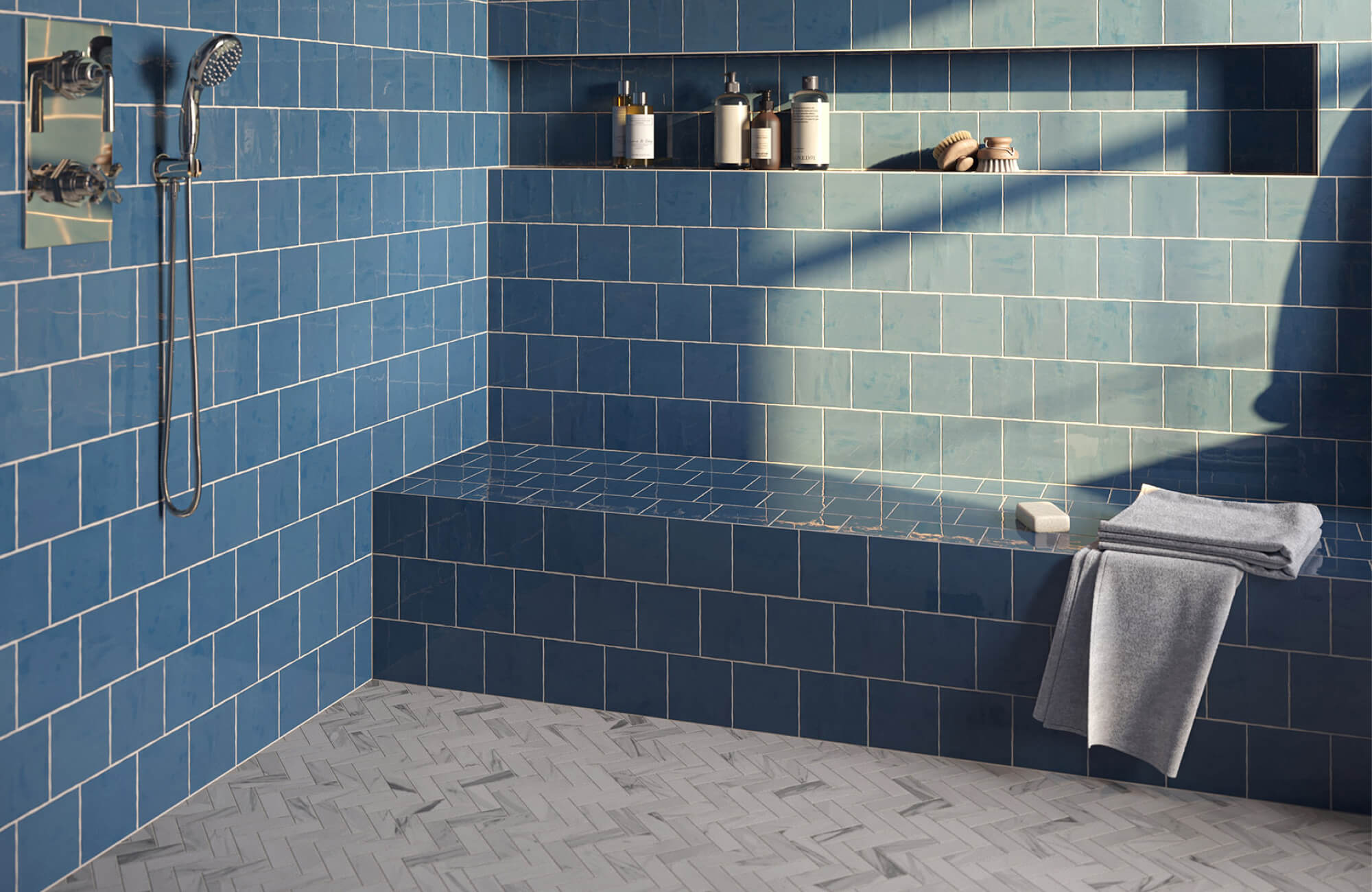 Blue tiled walk-in shower with a built-in bench, recessed wall niche, chrome shower fixtures, folded gray towels, and light gray herringbone mosaic floor tile.
