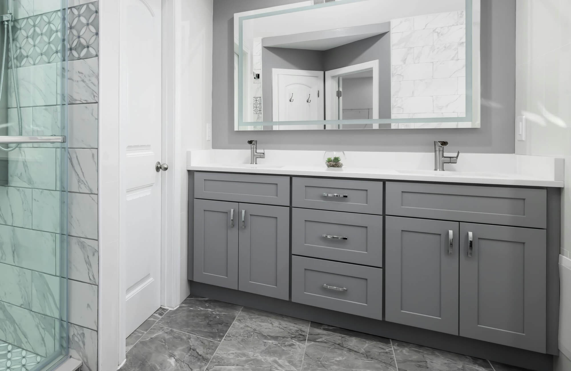 Modern bathroom with gray shaker-style vanity, dual sinks, marble-look tile flooring, and a glass shower with decorative accent tiles.