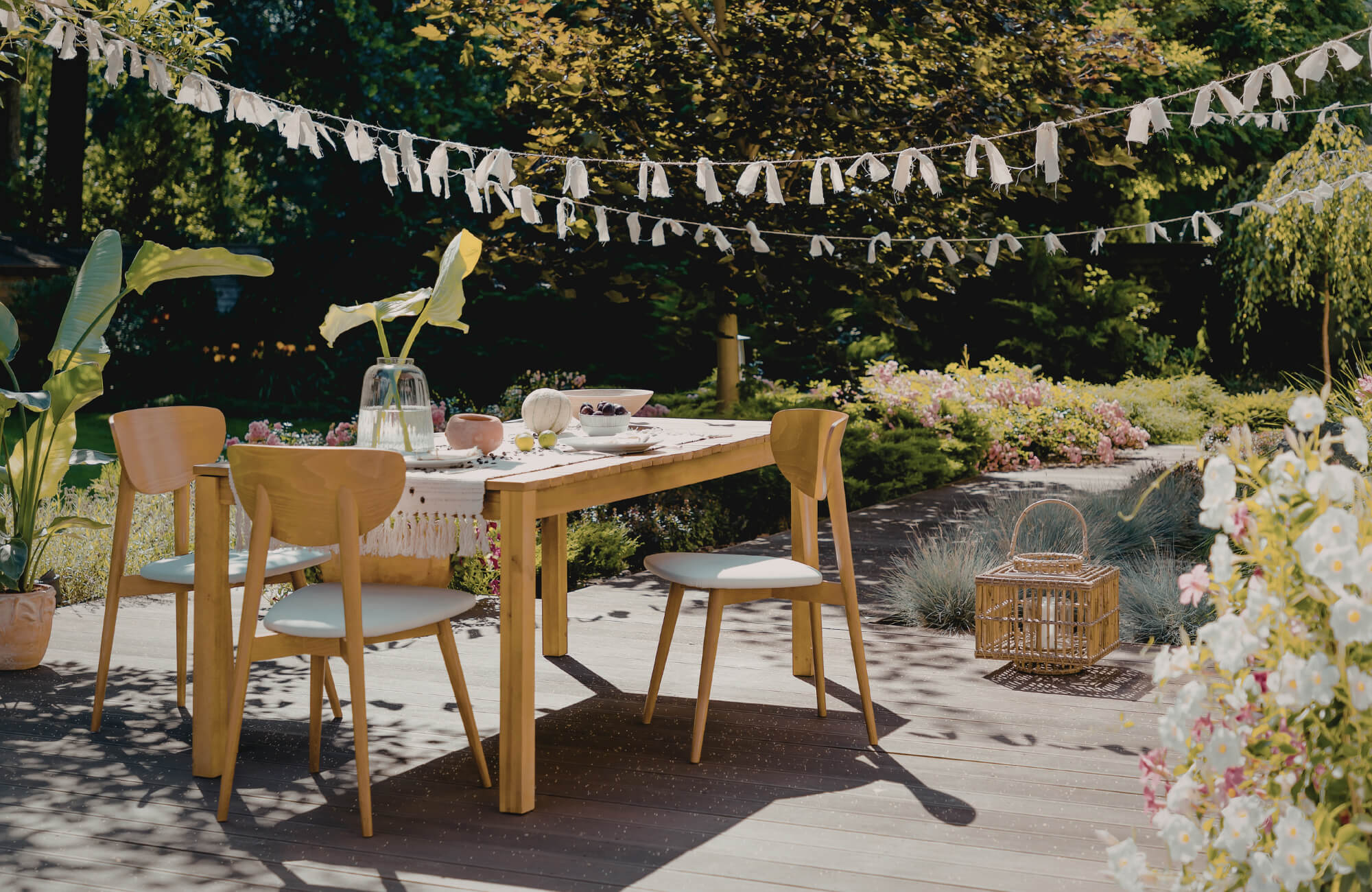 Outdoor wooden dining table with cushioned chairs on a garden deck, styled for relaxed dining and casual lounge-style entertaining.