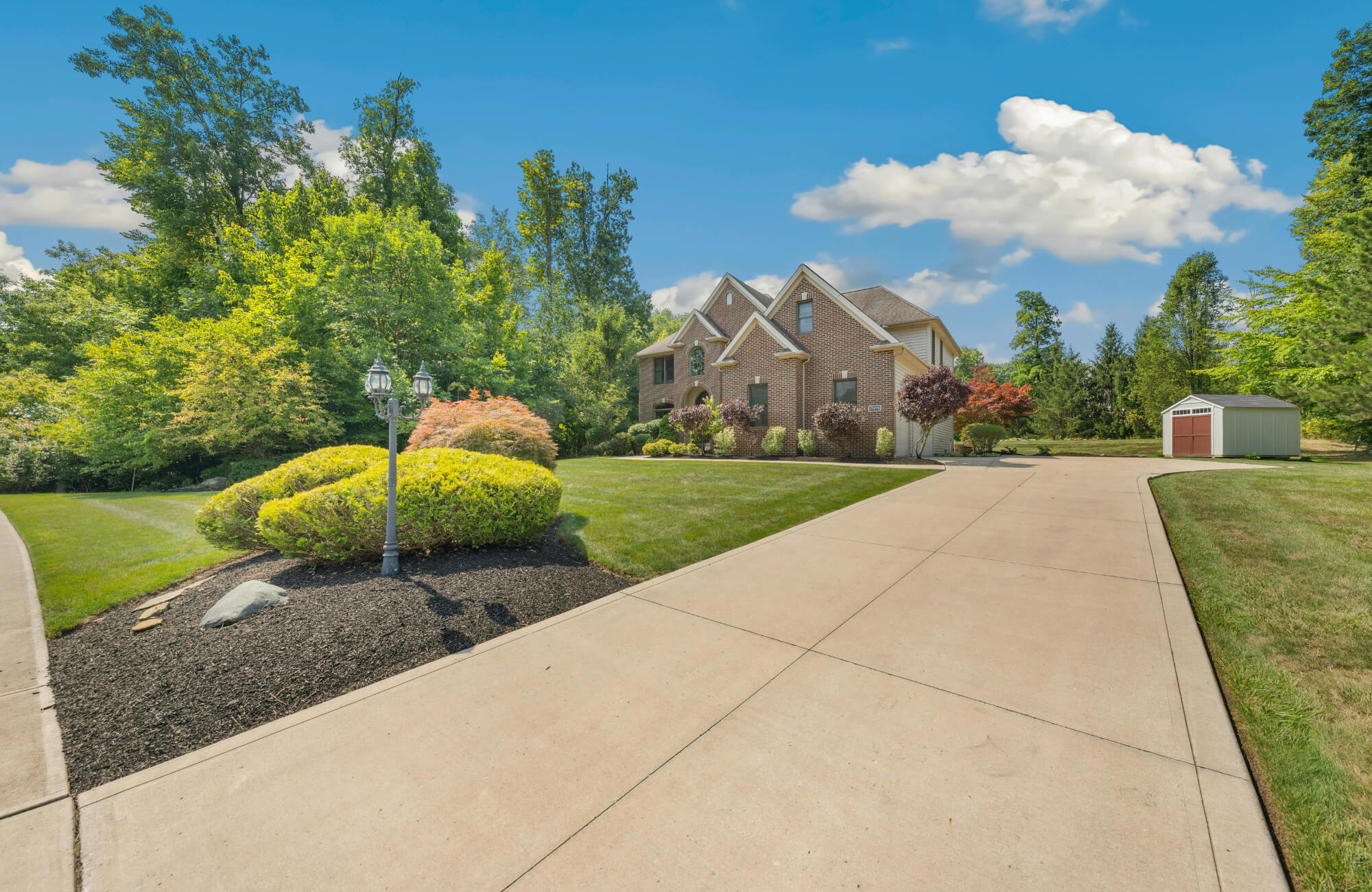 Large brick home with manicured landscaping, lush green lawn, and a wide concrete driveway under a bright blue sky with scattered clouds.