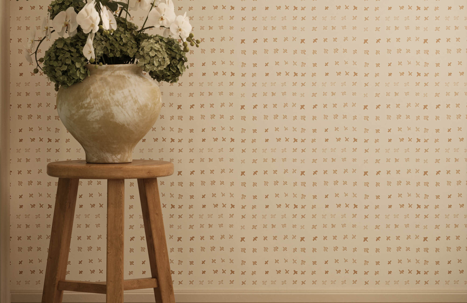 Elegant cream wallpaper with metallic cross motifs beside a wooden stool topped with white orchid arrangements.