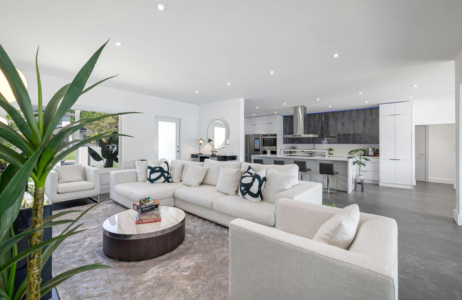 Modern open-concept living room with neutral sectional sofa and patterned pillows, styled with plants, marble coffee table, and sleek kitchen backdrop.