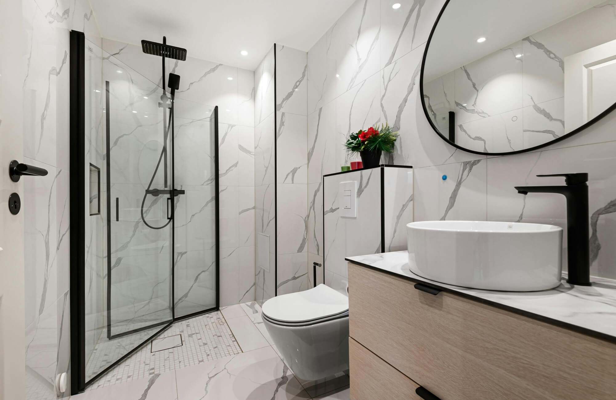 Modern minimalist bathroom with a black-framed oval mirror, walk-in shower with glass doors, and white marble-effect walls.