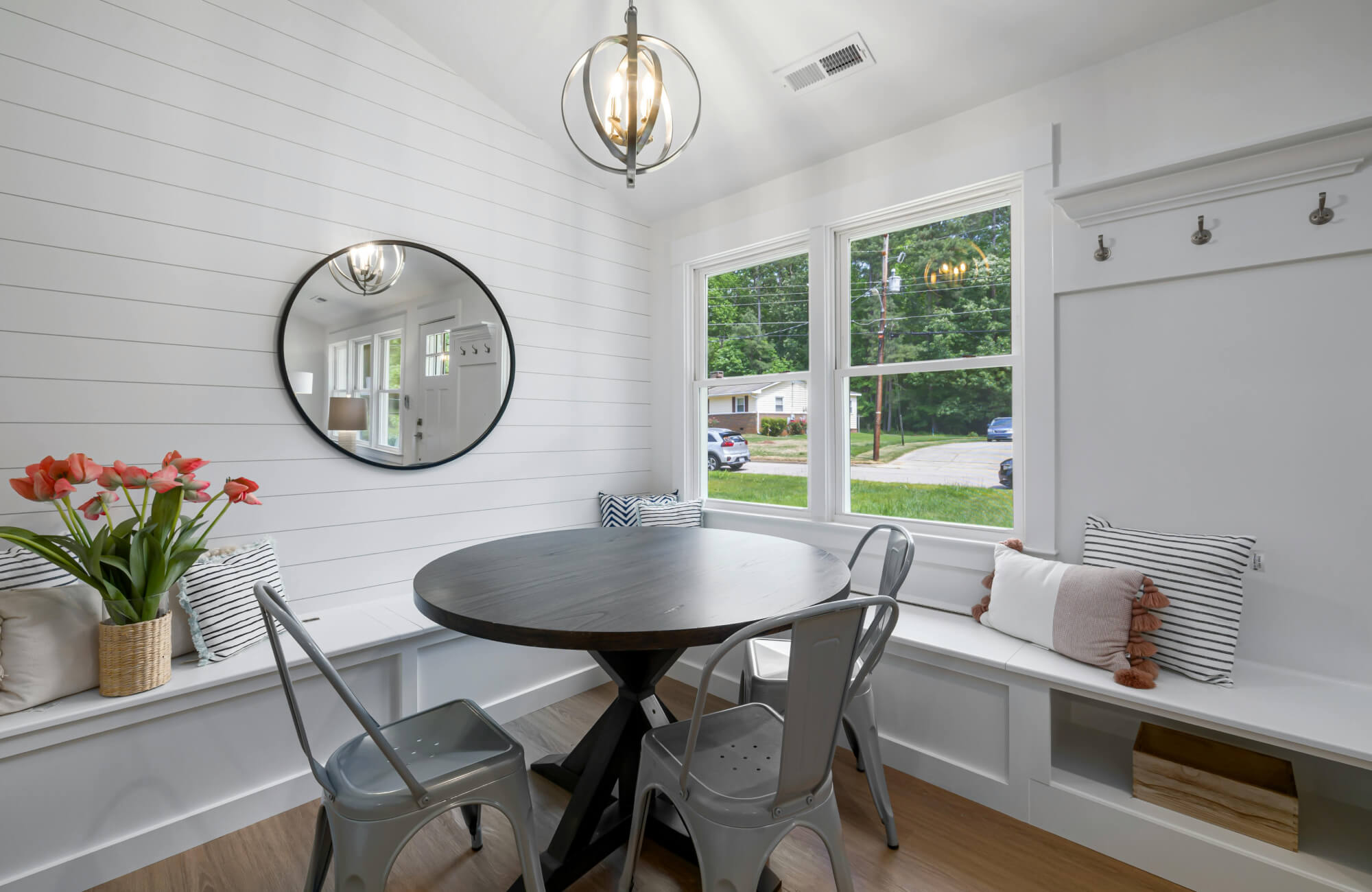 Bright and cozy breakfast nook with shiplap walls, built-in bench seating, a round black pedestal table, metal chairs, and a round mirror.